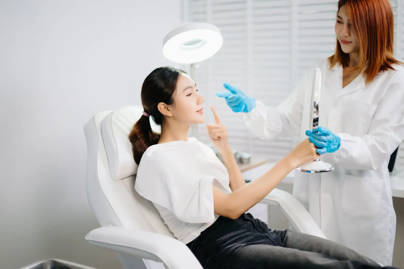Facelift 1 Woman in clinic chair uses mirror, guided by clinician in white coat and blue gloves, bright room.